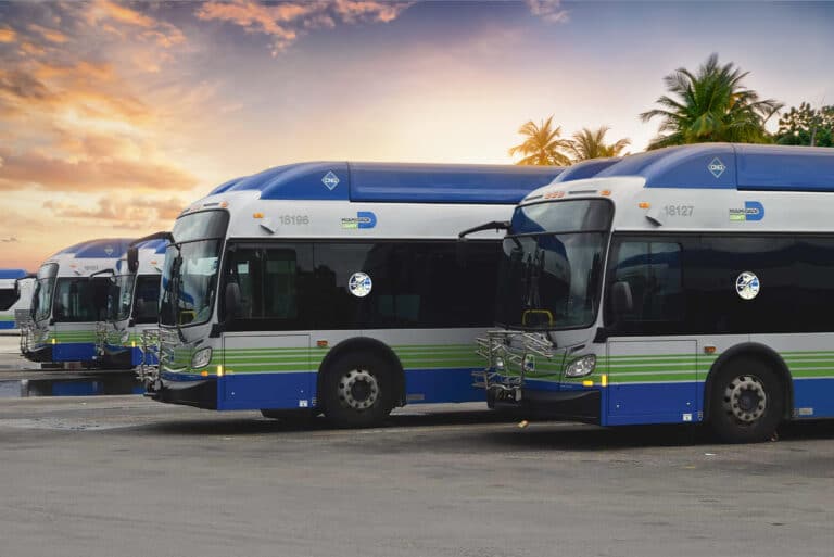 Miami-Dade CNG transit buses rest in a parking lot while the sun sets.
